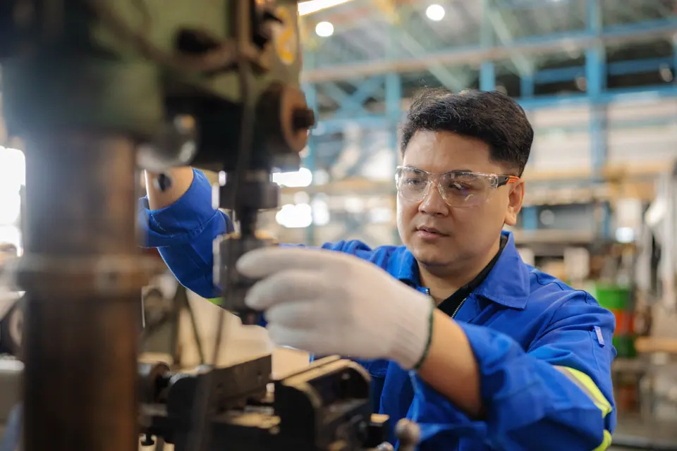 A blue-collar technician in protective gear inspects a power drill in a factory workshop