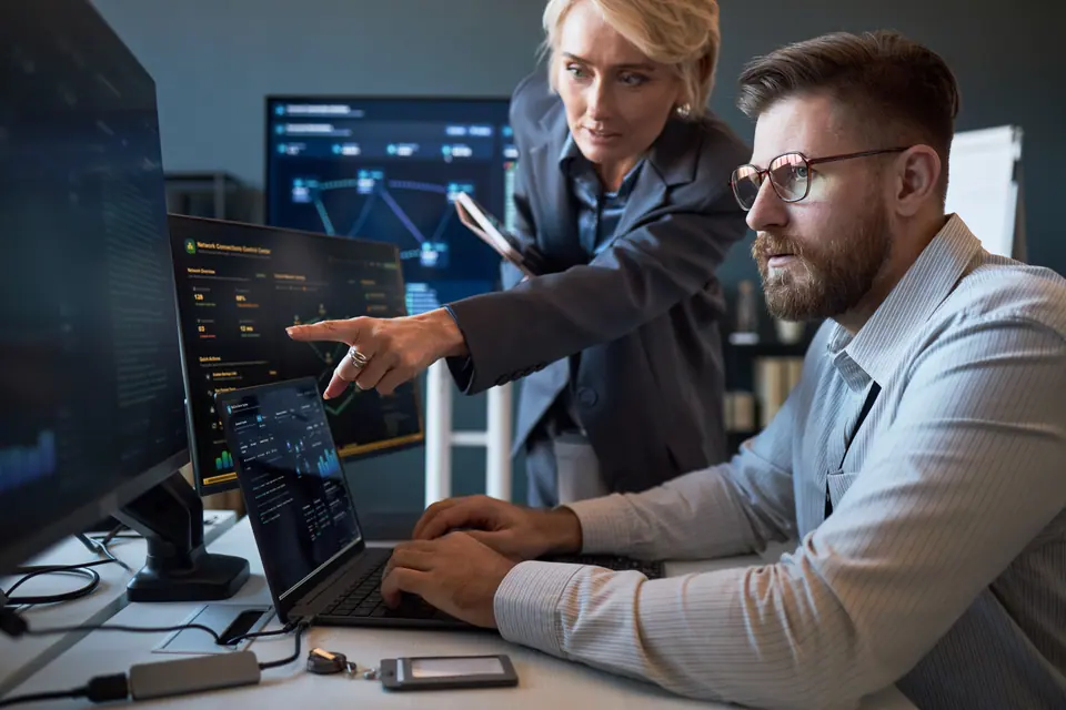 Caucasian businesswoman pointing at cybersecurity dashboard while Caucasian businessman analyzing data on laptop and monitors