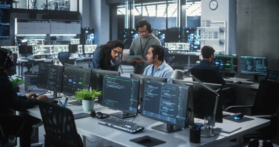Group of three young indian software engineers use computer to discuss a technological project in modern industrial office