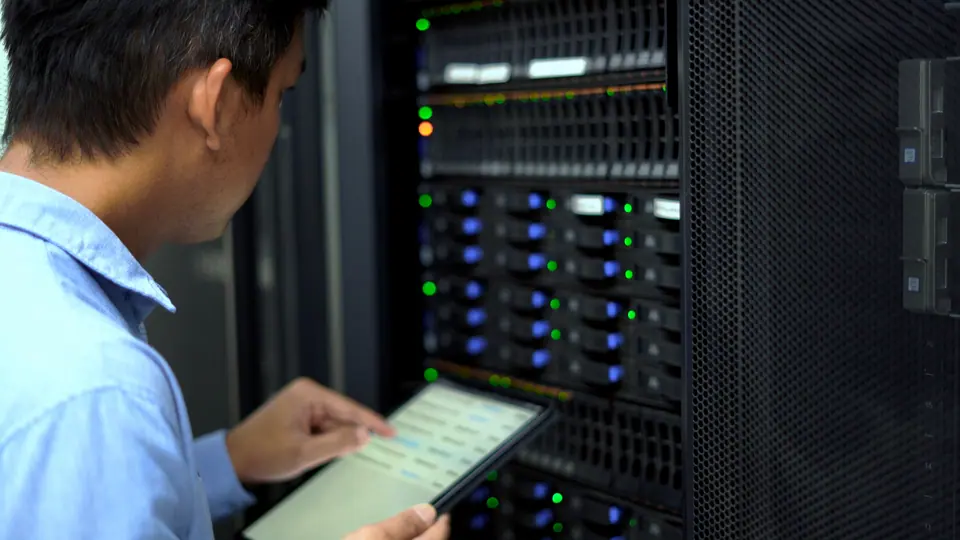 An IT technician inspects and manages a server rack in a data center using a tablet, representing cloud computing, cybersecurity, and network…