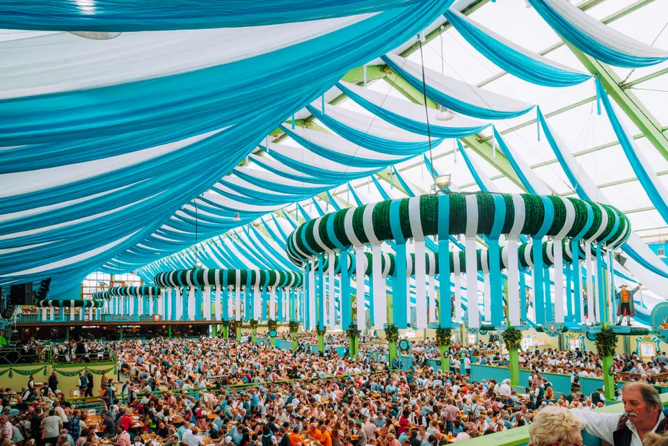Large crowd gathers under blue-and-white draped ceiling inside a Munich Volksfest beer tent in Bavaria, Germany, enjoying a lively communal celebration…