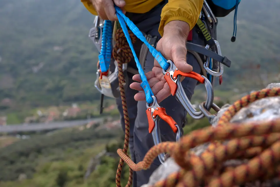 Mature male rock climber secures rope onto rock on cliff above valley and hills, moody sky, Liguria