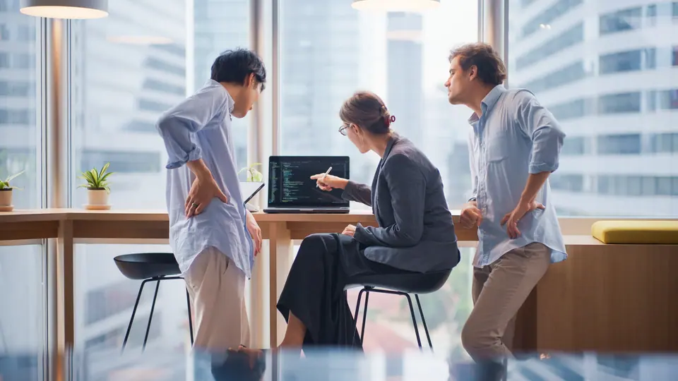 Three software engineers, including two men and one woman, are gathered around a laptop discussing programming code in a modern, high-rise office with…