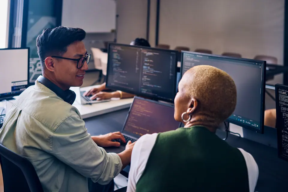 Young computer programmer concentrating on dual monitors, developing software or analyzing data in a busy office environment